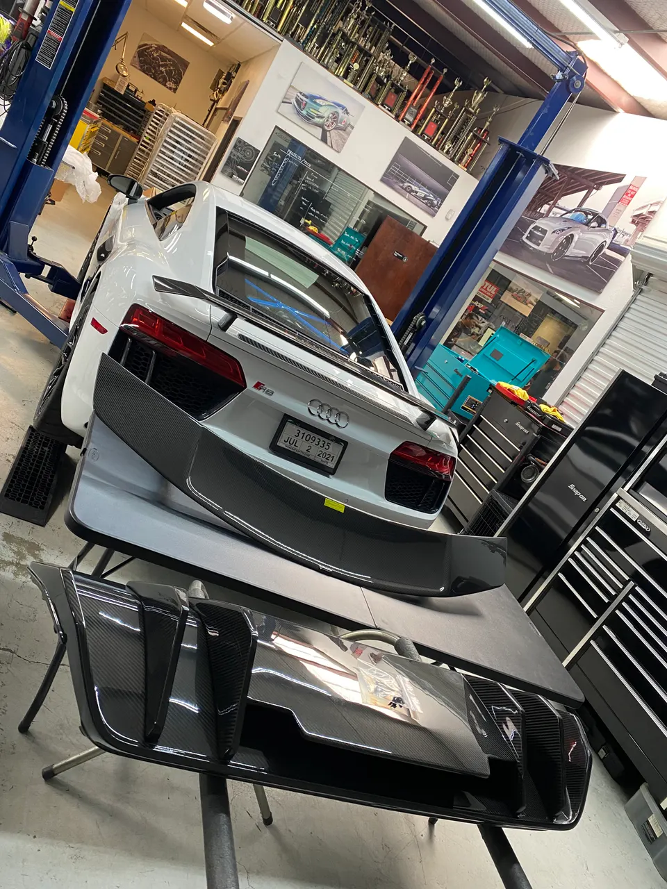 A white Audi R8 on a lift in a performance shop with carbon fiber aerodynamic parts, including a rear diffuser and spoiler, on a table.