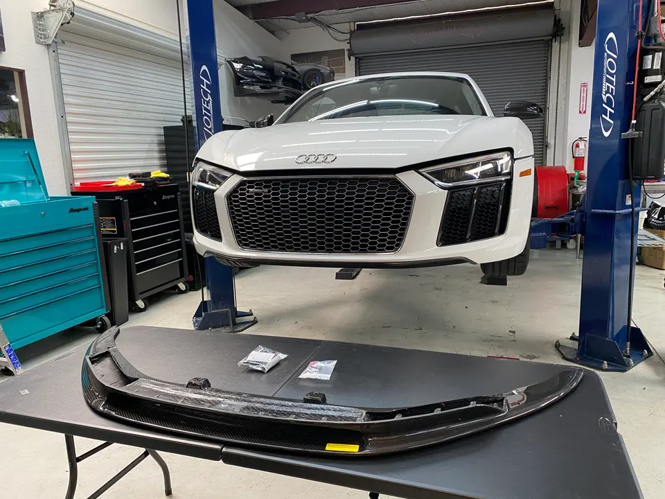 A white Audi R8 sports car is elevated on a lift in a garage, with a new carbon fiber front splitter on a table in the foreground.