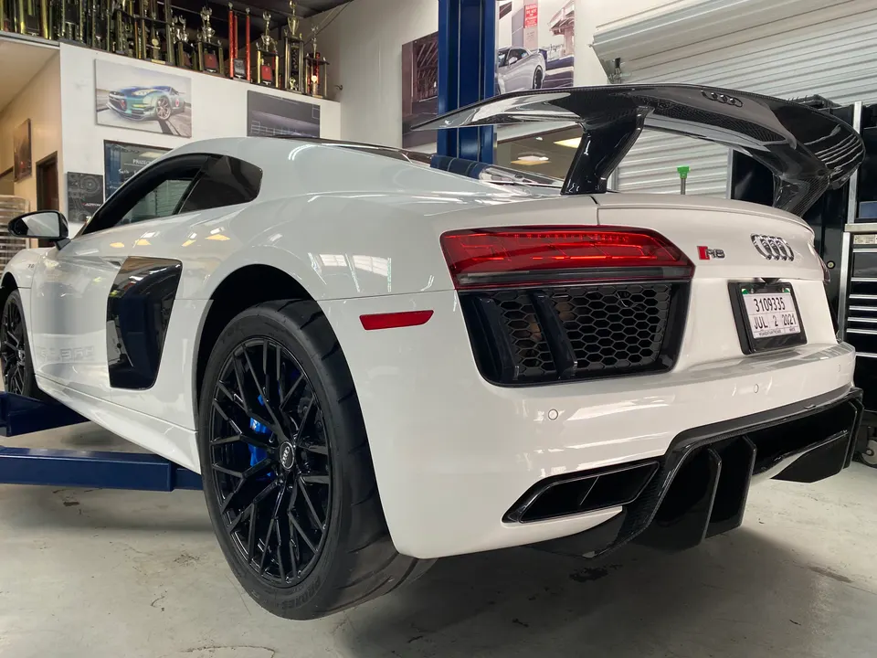 White Audi R8 on a lift in a performance shop, showcasing its carbon fiber wing, diffuser, and black aftermarket wheels.
