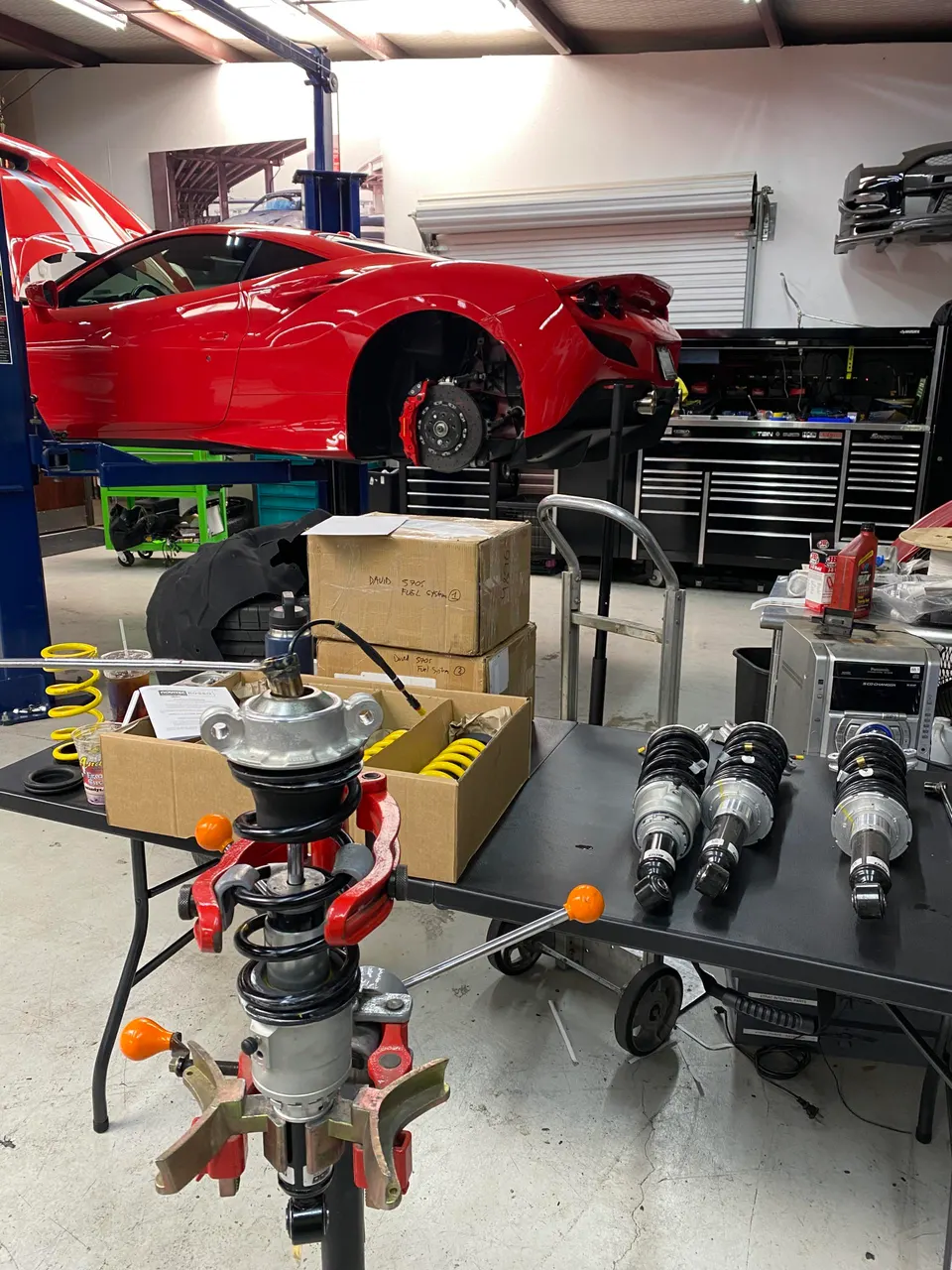A red Ferrari sports car on a lift in a performance tuning shop, with suspension components and tools on a table.
