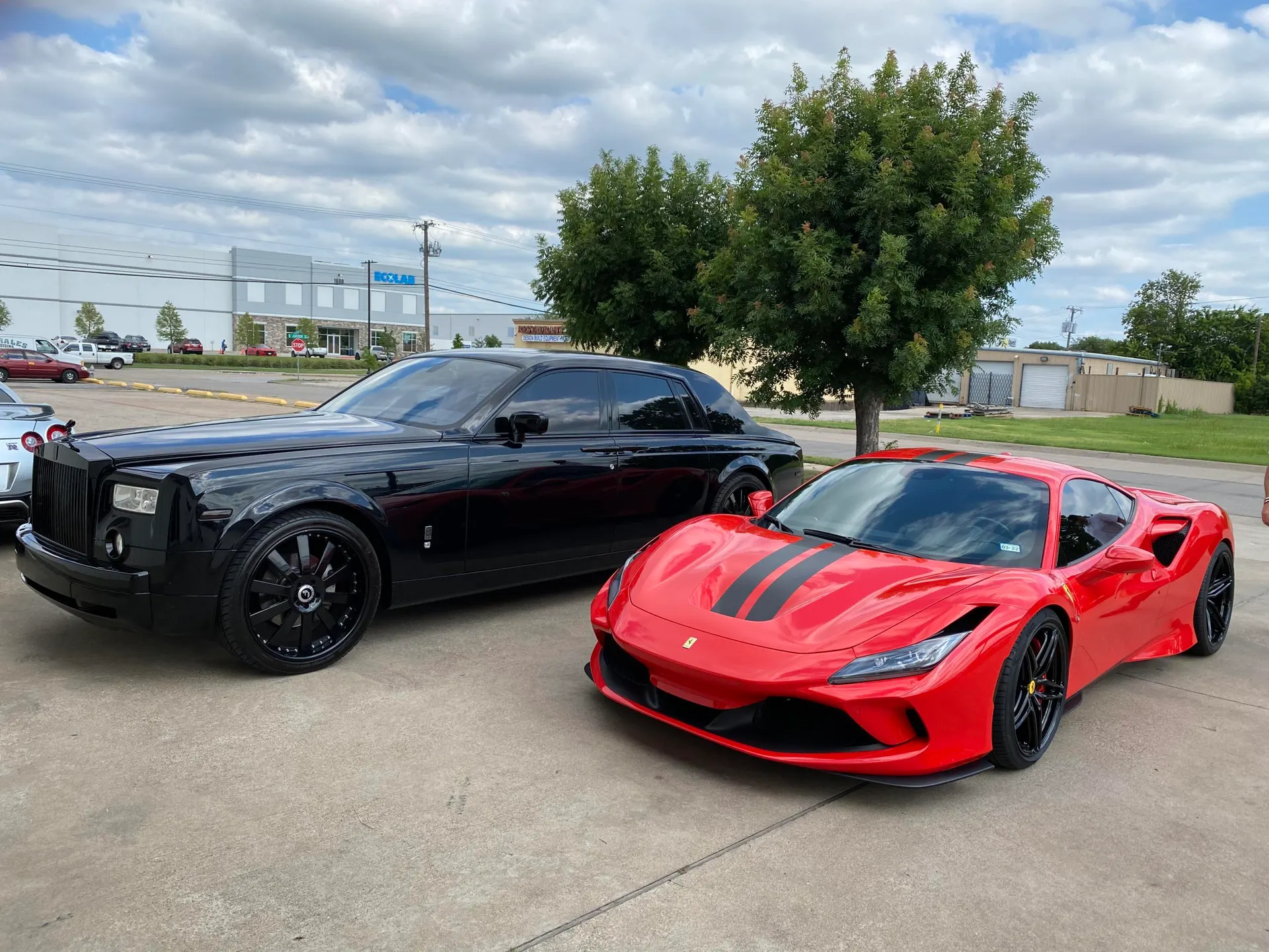 A black Rolls-Royce Phantom parked next to a red Ferrari F8 Tributo with black racing stripes in an outdoor lot.