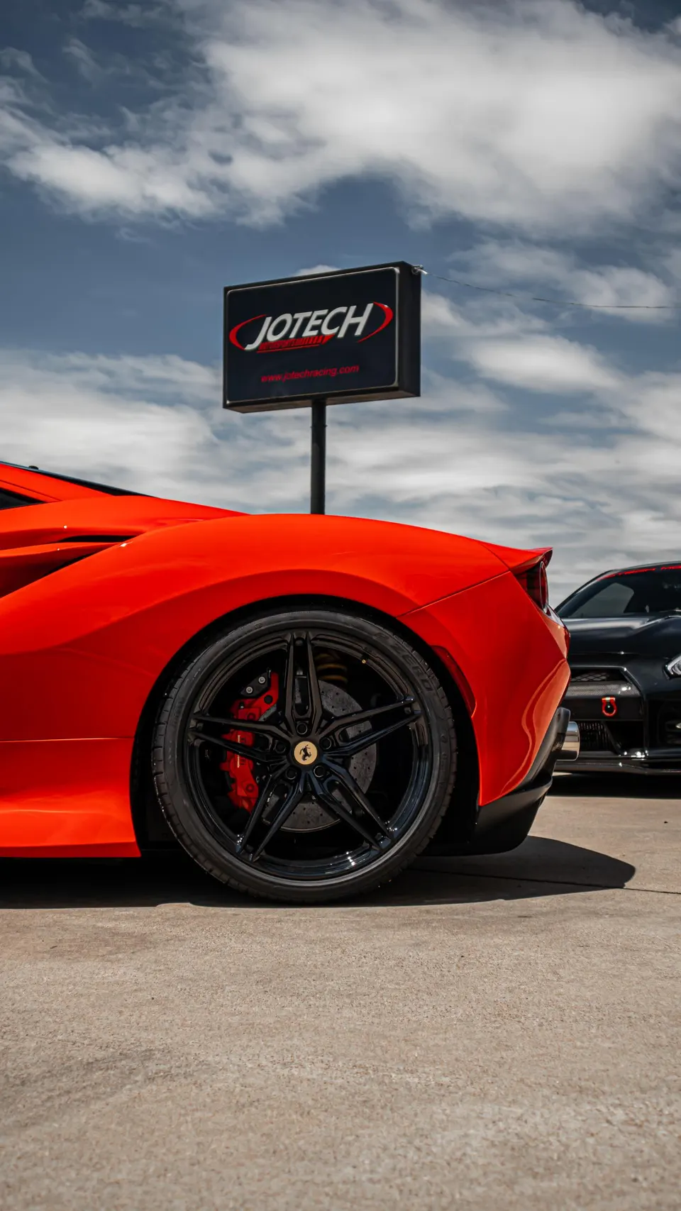 Rear view of a red Ferrari with black wheels and red calipers, parked next to a black Nissan GT-R outside Jotech Motorsports.
