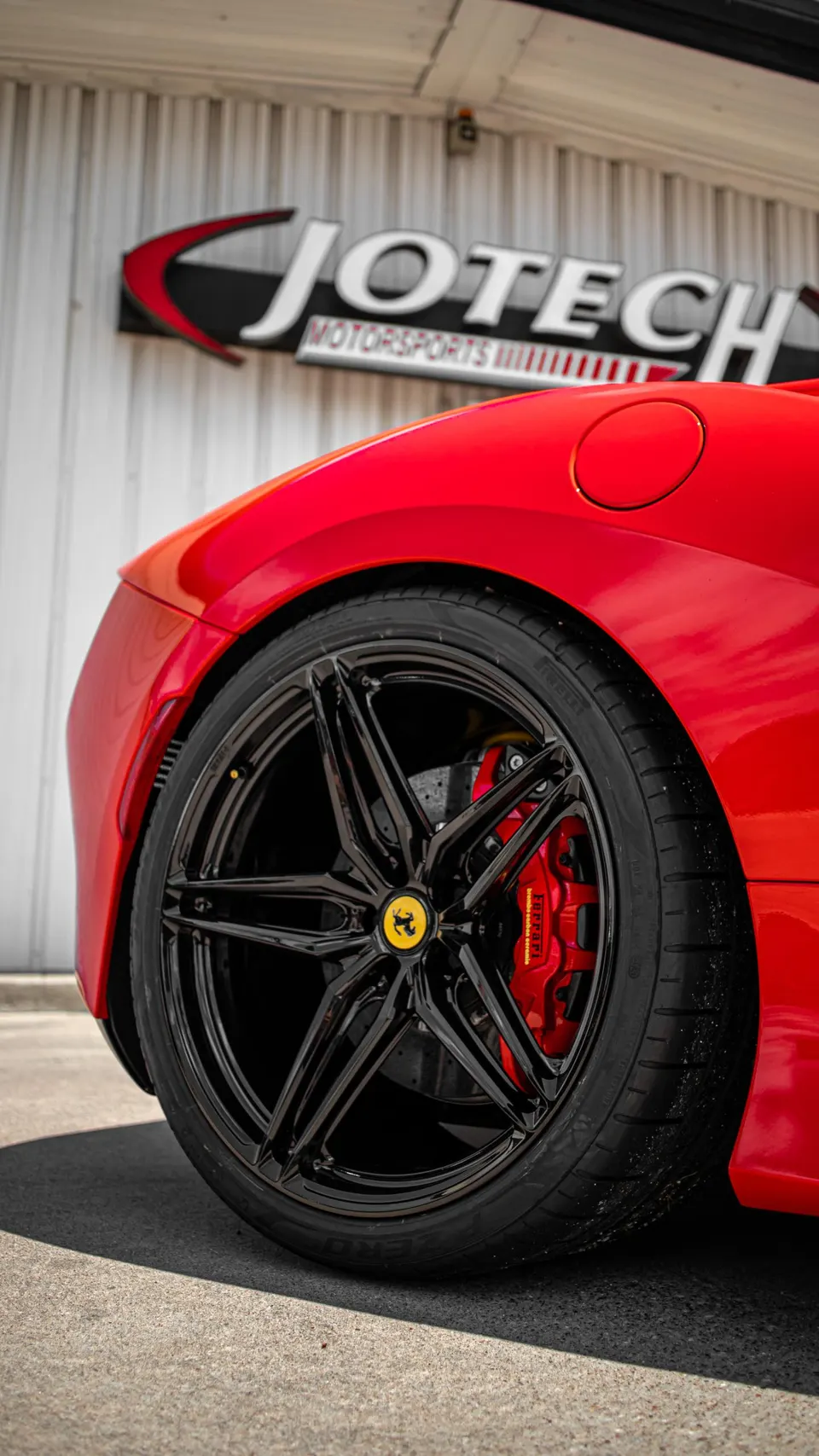 Close-up of a red Ferrari's black multi-spoke wheel, red brake caliper, and Pirelli tire in front of Jotech Motorsports.