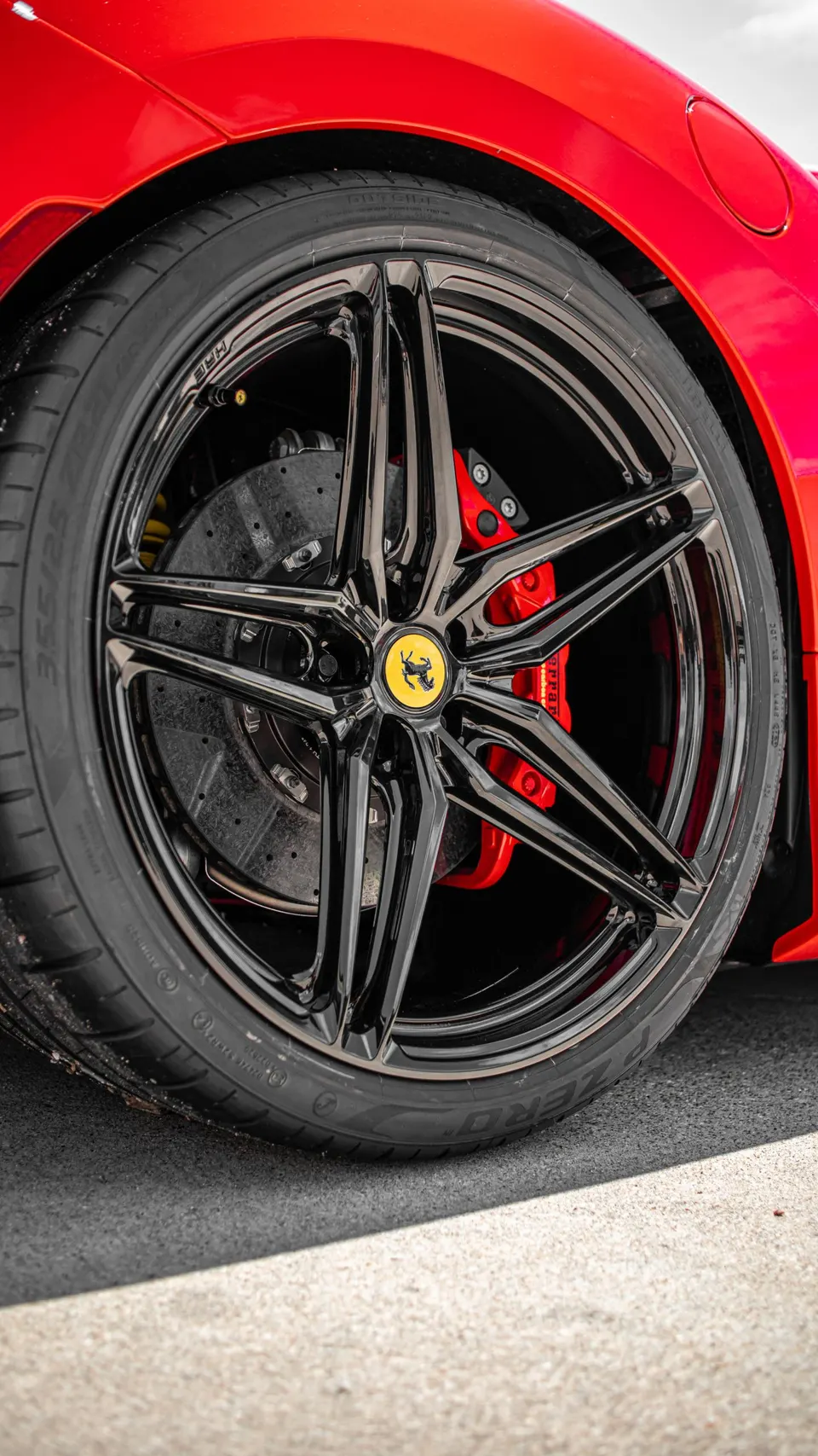 Close-up of a red Ferrari's black multi-spoke wheel with yellow center cap, red brake calipers, and carbon ceramic rotors.