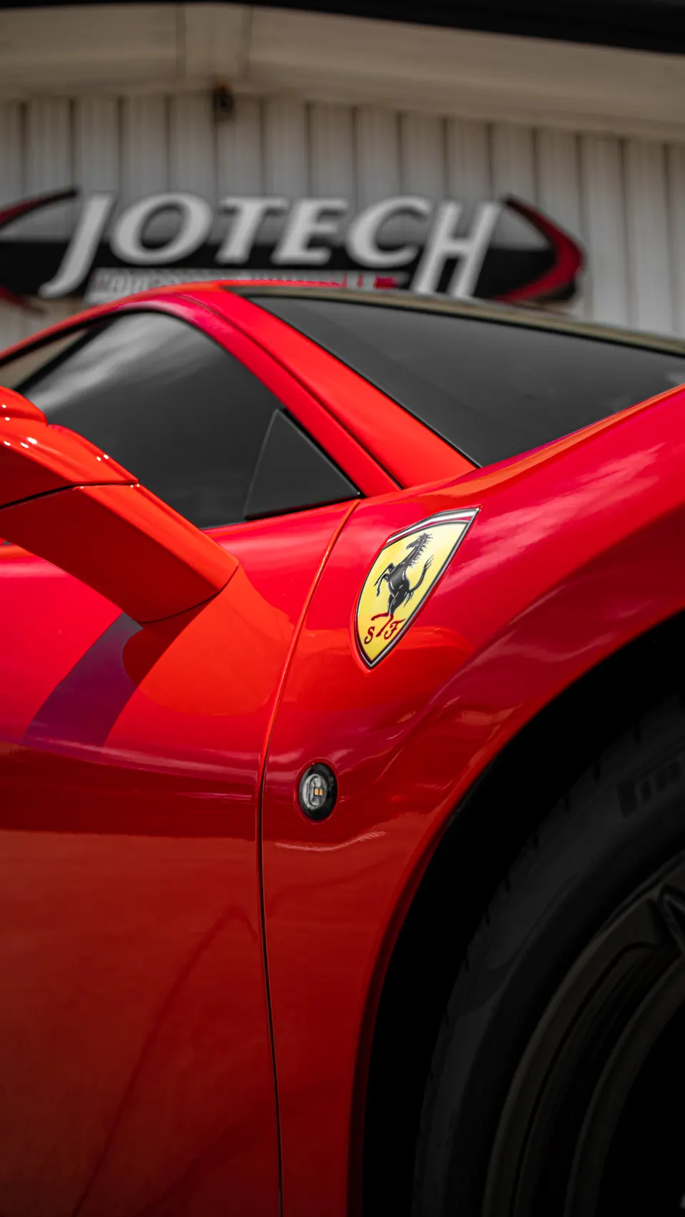 Close-up of a red Ferrari's fender with the iconic prancing horse badge, parked in front of a Jotech sign.