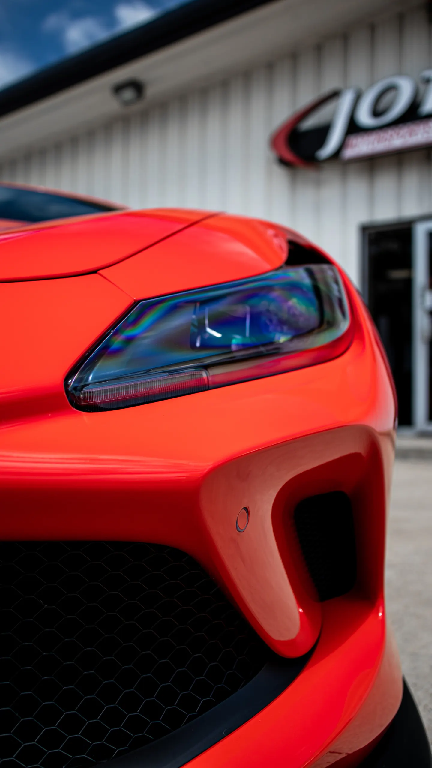Close-up of the front of a red sports car, showing the headlight, bumper, and grille, with a Jotech Motorsports sign in the background.