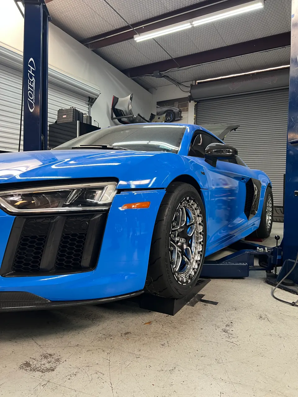 A blue Audi R8 on a lift in a performance tuning shop, featuring aftermarket drag wheels and performance tires.