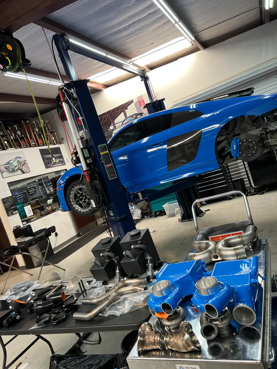 A blue Audi R8 on a lift in a performance shop with a turbo kit and other aftermarket parts laid out on tables.