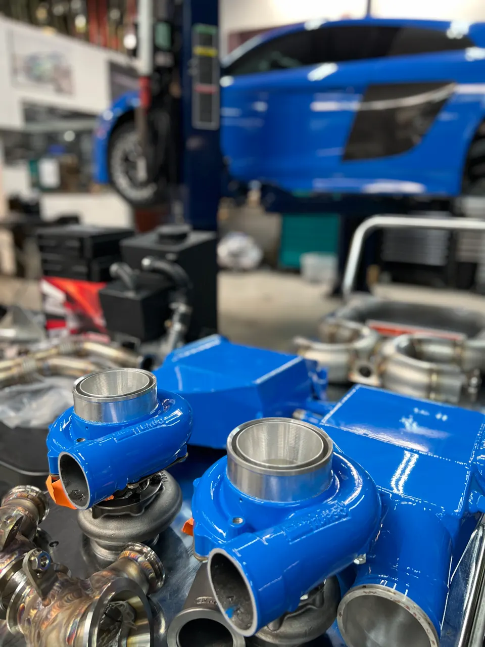 Blue turbochargers and boost piping components laid out in a performance tuning shop with an Audi R8 on a lift.