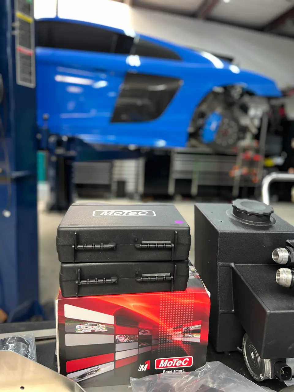 MoTeC engine management systems and a custom tank on a workbench in a performance tuning shop with a blue Audi R8 on a lift.