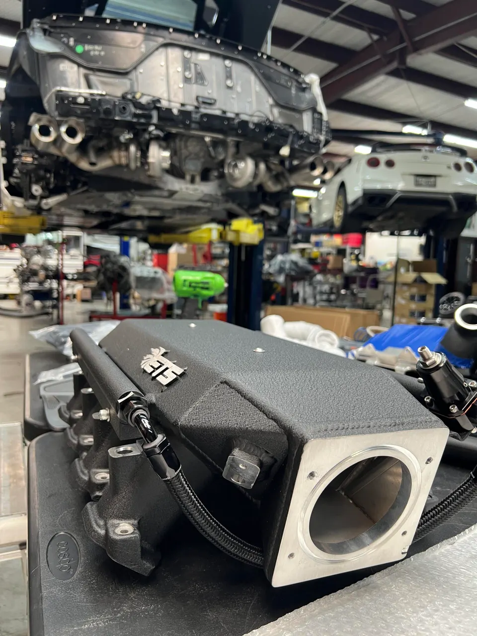 A black aftermarket intake manifold with a fuel rail on a workbench in a performance tuning shop with cars on lifts.