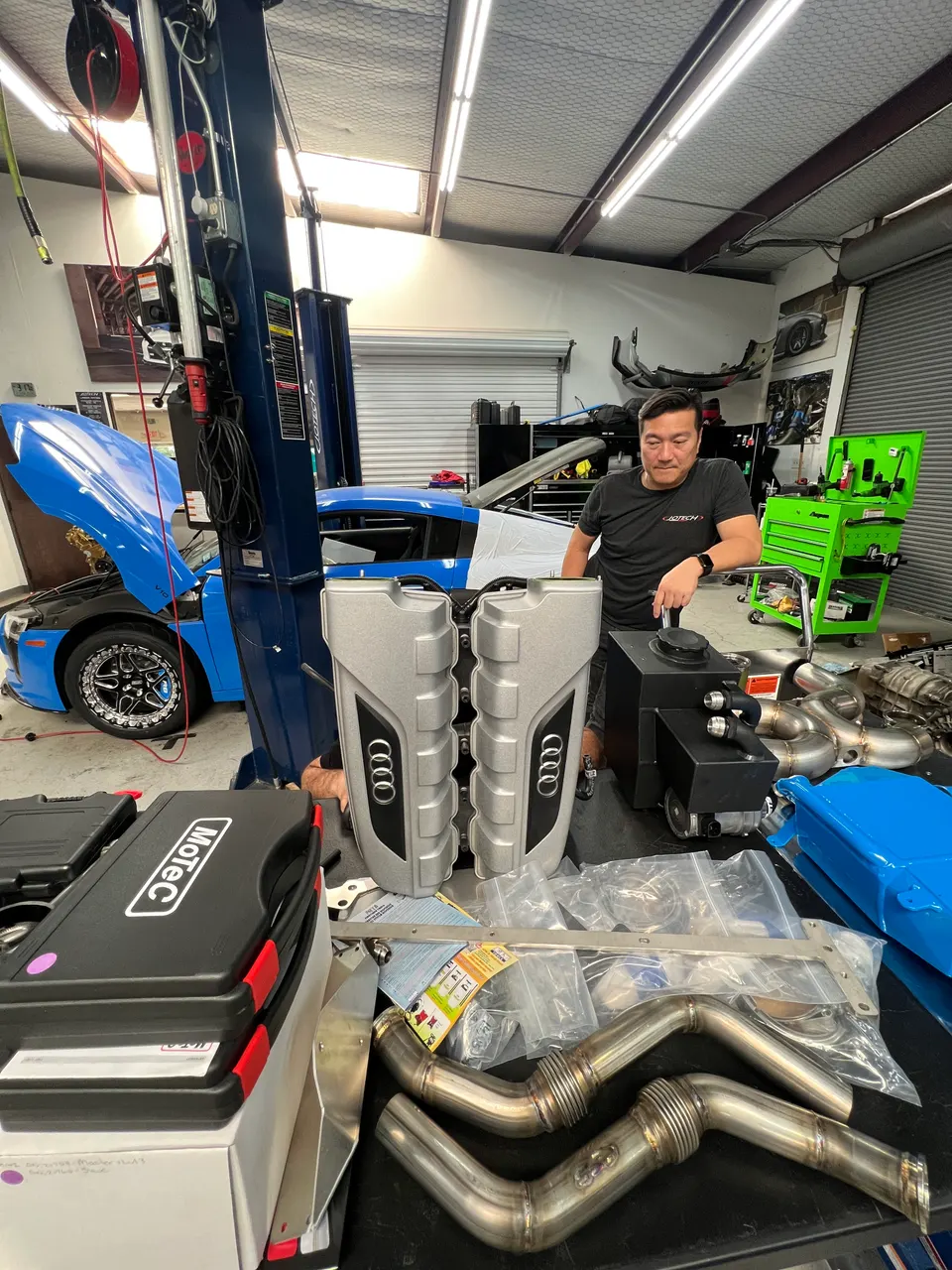 Jotech Motorsports mechanic with Audi R8 performance parts: MoTeC system, exhaust, and valve covers, in a shop with a blue R8.