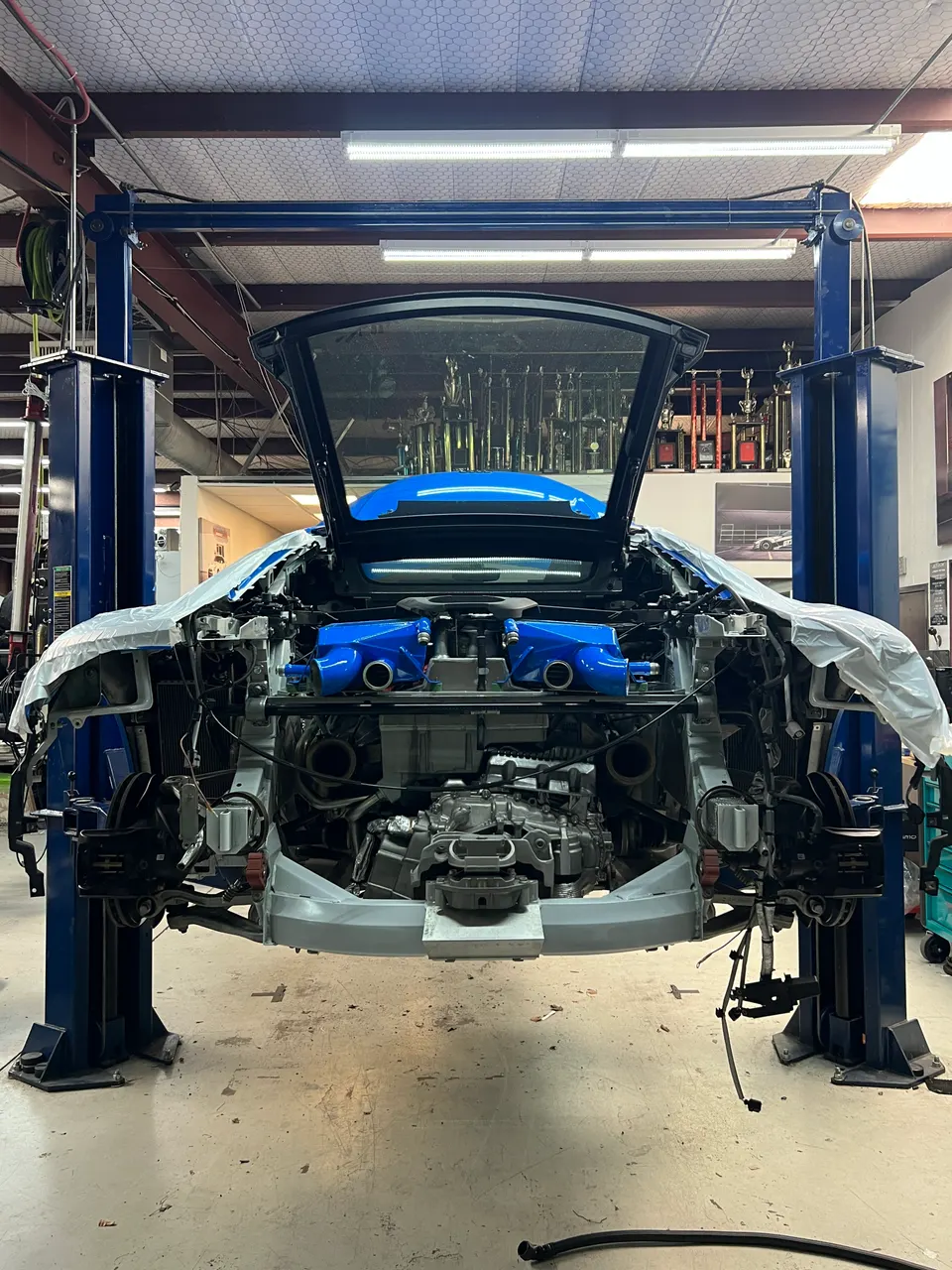 Rear view of a Lamborghini Huracan on a lift in a shop, undergoing disassembly with the engine and blue induction system exposed.