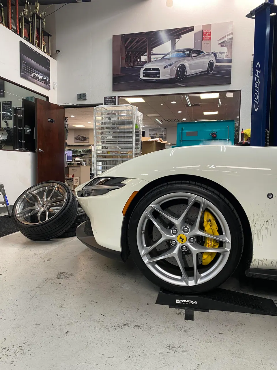 A white Ferrari with yellow brake calipers and a silver wheel on a ramp in a performance tuning shop.