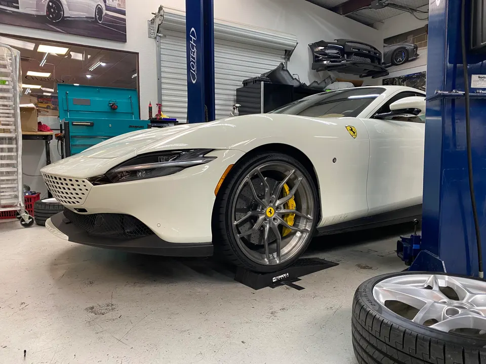 A white Ferrari Roma with aftermarket wheels and yellow brake calipers in a Jotech Motorsports shop, with a spare wheel in the foreground.