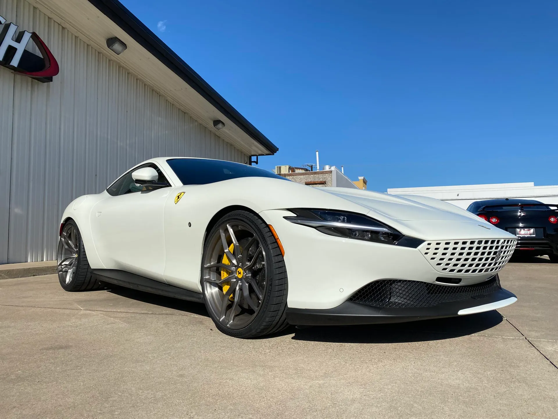 A white Ferrari Roma with custom wheels and yellow brake calipers parked outside a Jotech Motorsports facility.