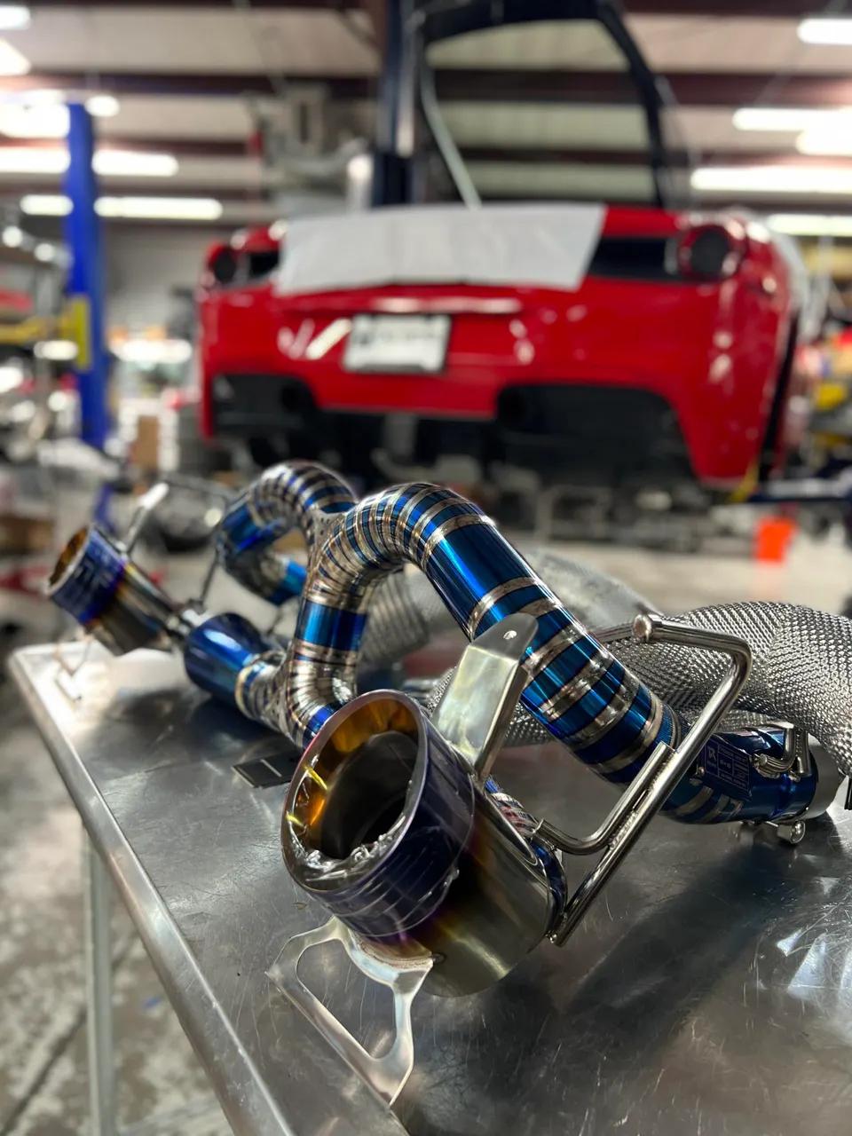 A blue titanium aftermarket exhaust system on a workbench in a performance tuning shop, with a red Ferrari in the background.