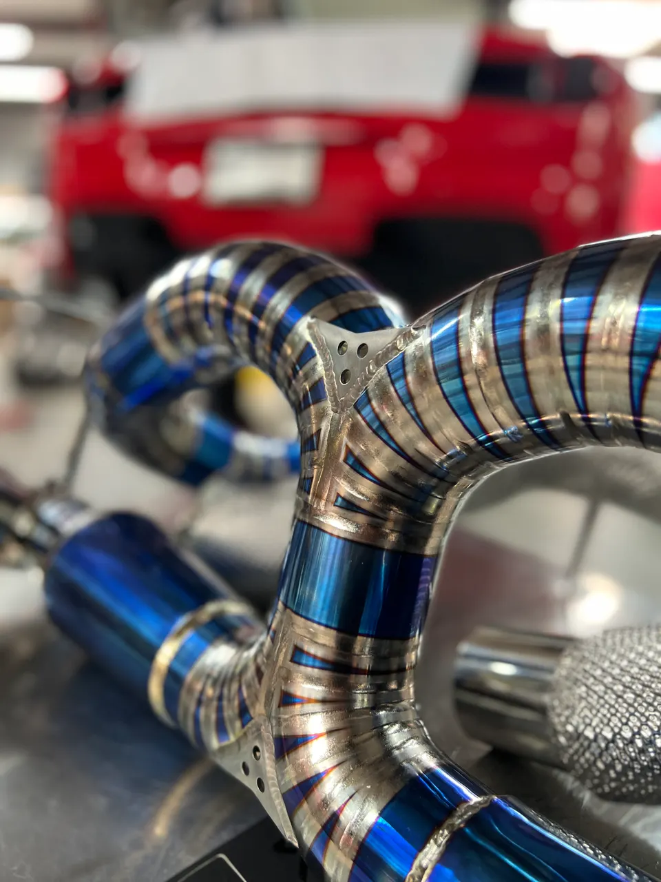 A close-up of a custom titanium exhaust system with blue heat-treated sections and intricate welds, with a red GT-R in the background.