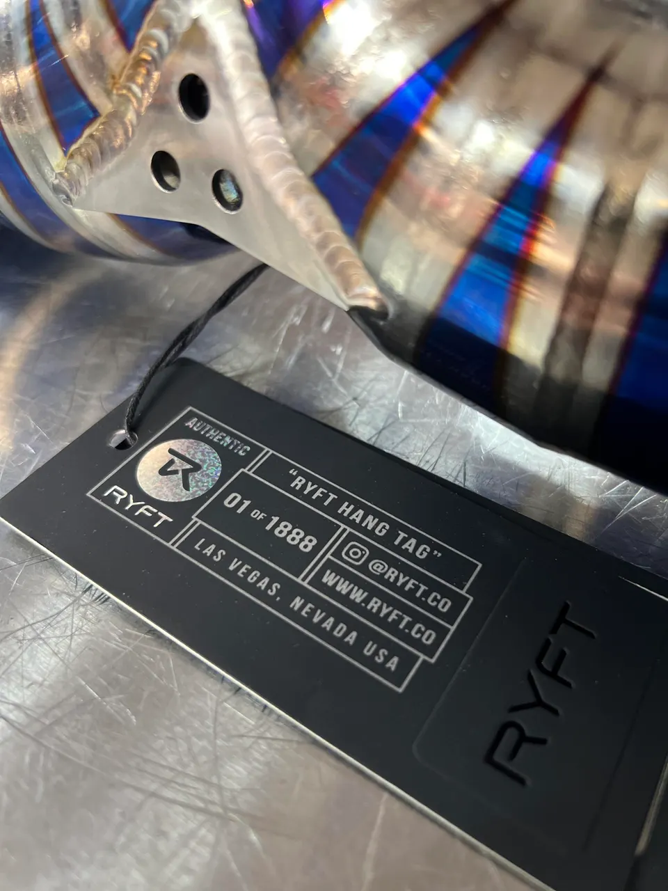 Close-up of a RYFT titanium exhaust component with heat coloring and a branded hang tag.