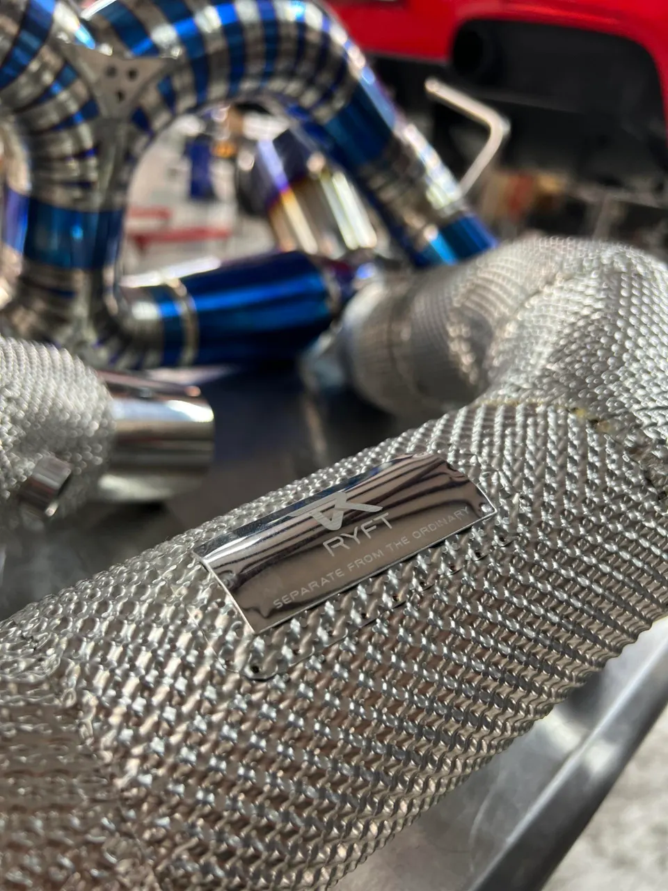 Close-up of a RYFT performance exhaust system with heat wrap and titanium piping in a shop setting.