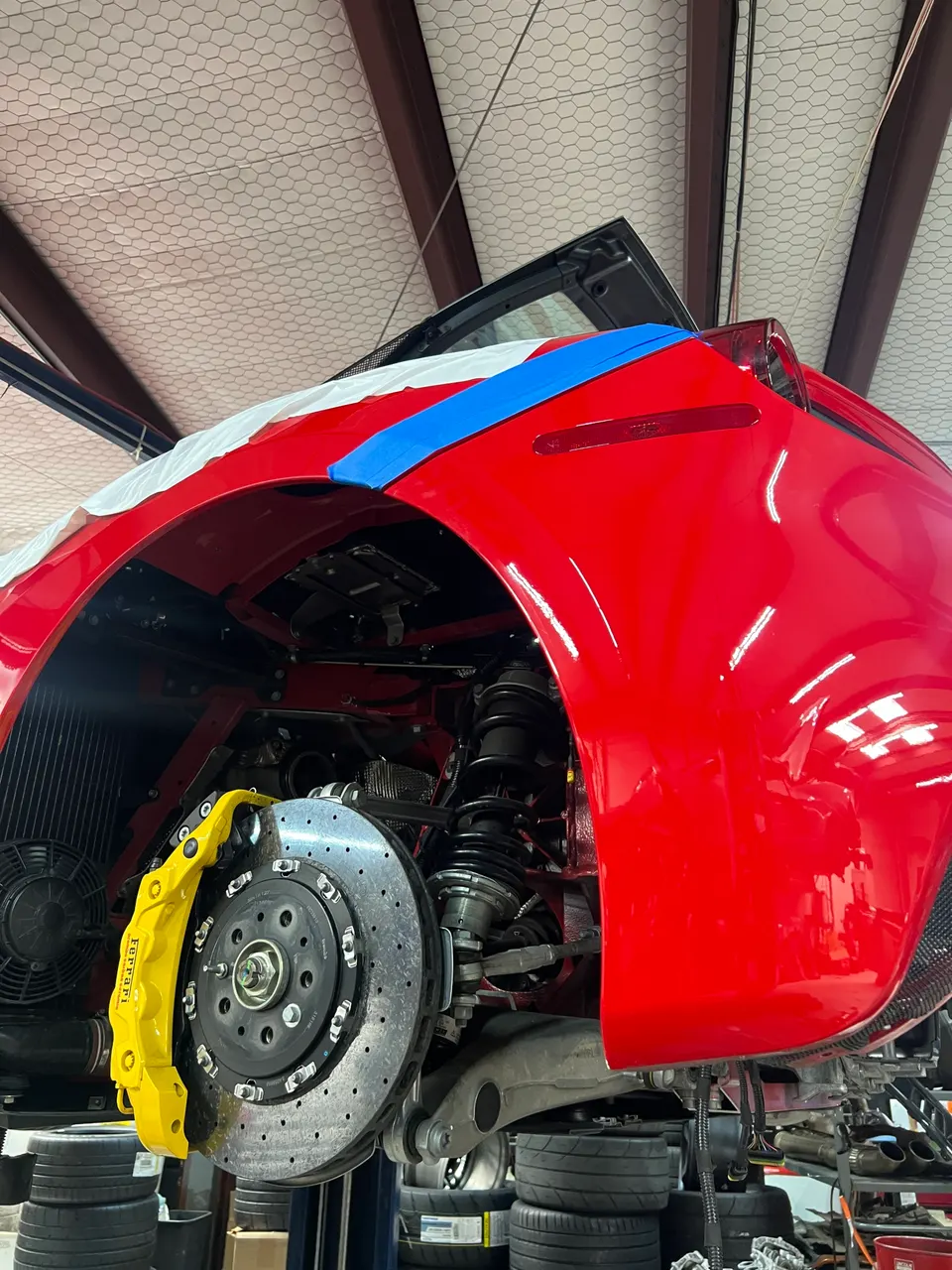 Red Ferrari on a lift in a shop, showcasing the yellow brake caliper and drilled rotor.