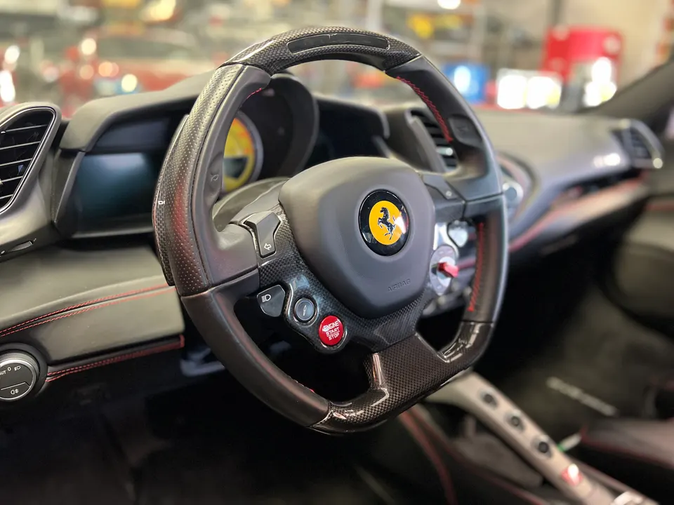 Close-up of a Ferrari steering wheel with carbon fiber trim and red stitching, showing the dashboard and controls.