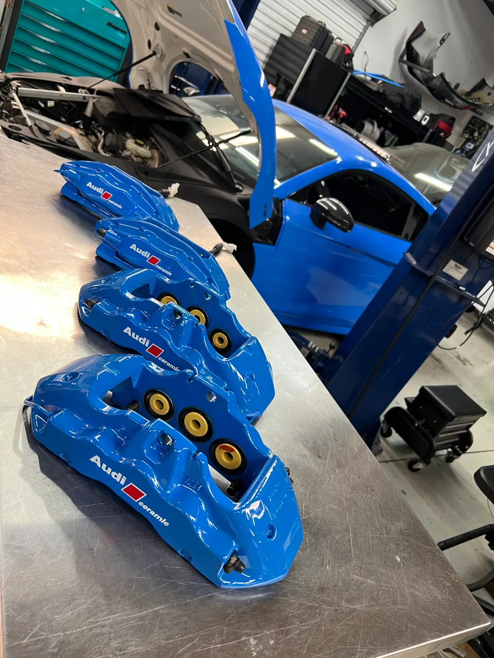Blue Audi ceramic brake calipers on a workbench in a performance tuning shop, with an Audi R8 in the background.
