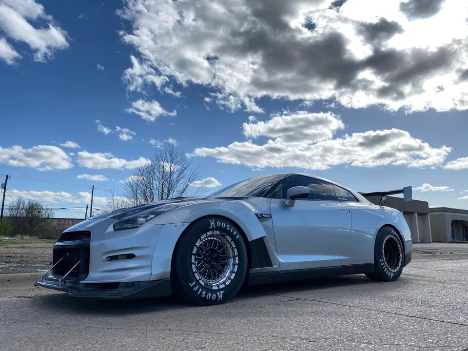 Silver Nissan GT-R drag car with Hoosier slicks and aggressive aero parked outdoors under a cloudy sky.