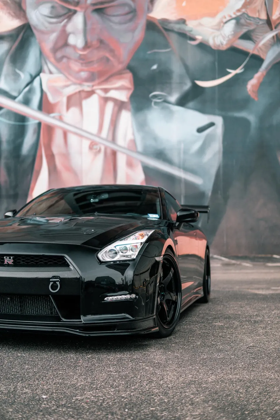 A black Nissan GT-R R35 with aftermarket wheels and a front tow hook parked in front of a colorful mural.