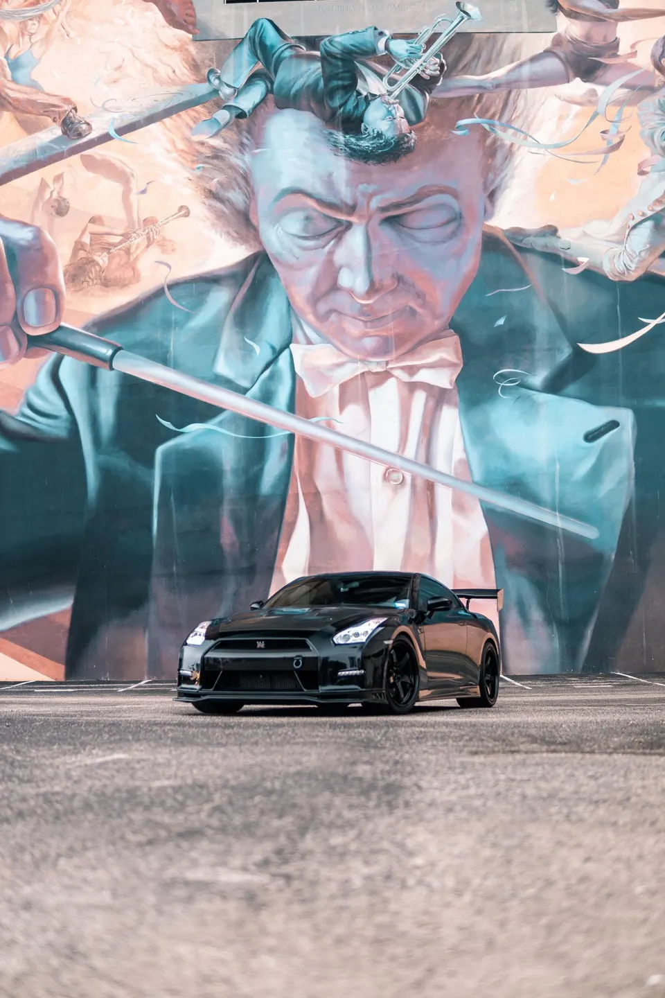 A black Nissan GT-R with aftermarket wheels and a rear wing parked in front of a large, colorful mural.