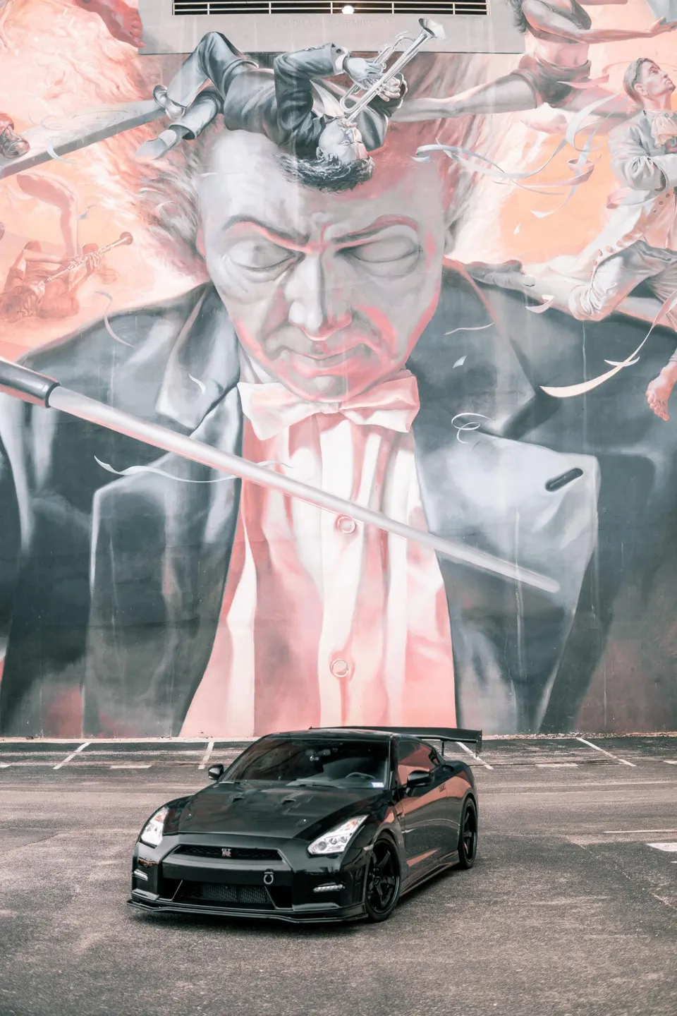 Black Nissan GT-R with aftermarket wheels and a large rear wing parked in front of a vibrant classical music mural.
