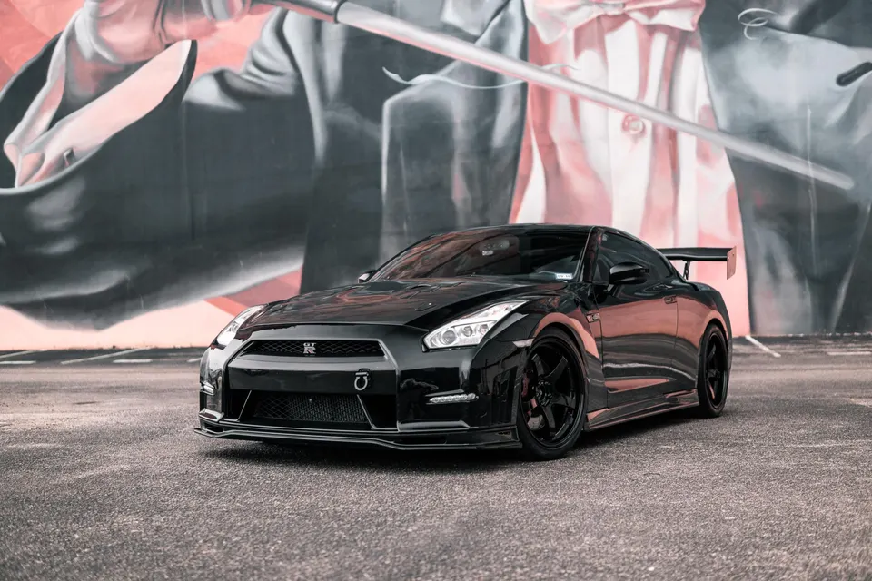Black Nissan GT-R with aftermarket wheels and aero kit parked in front of a colorful mural, showcasing its sleek design.