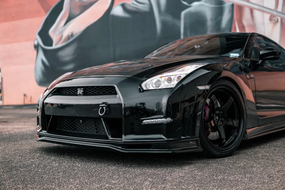 A black Nissan GT-R R35 with carbon fiber front splitter and aftermarket wheels parked in front of a mural.