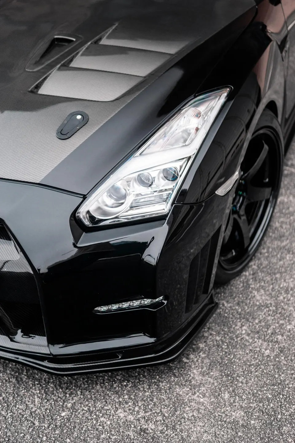 Close-up of a black Nissan GT-R's front end, showcasing its carbon fiber hood, custom bumper, and headlight.