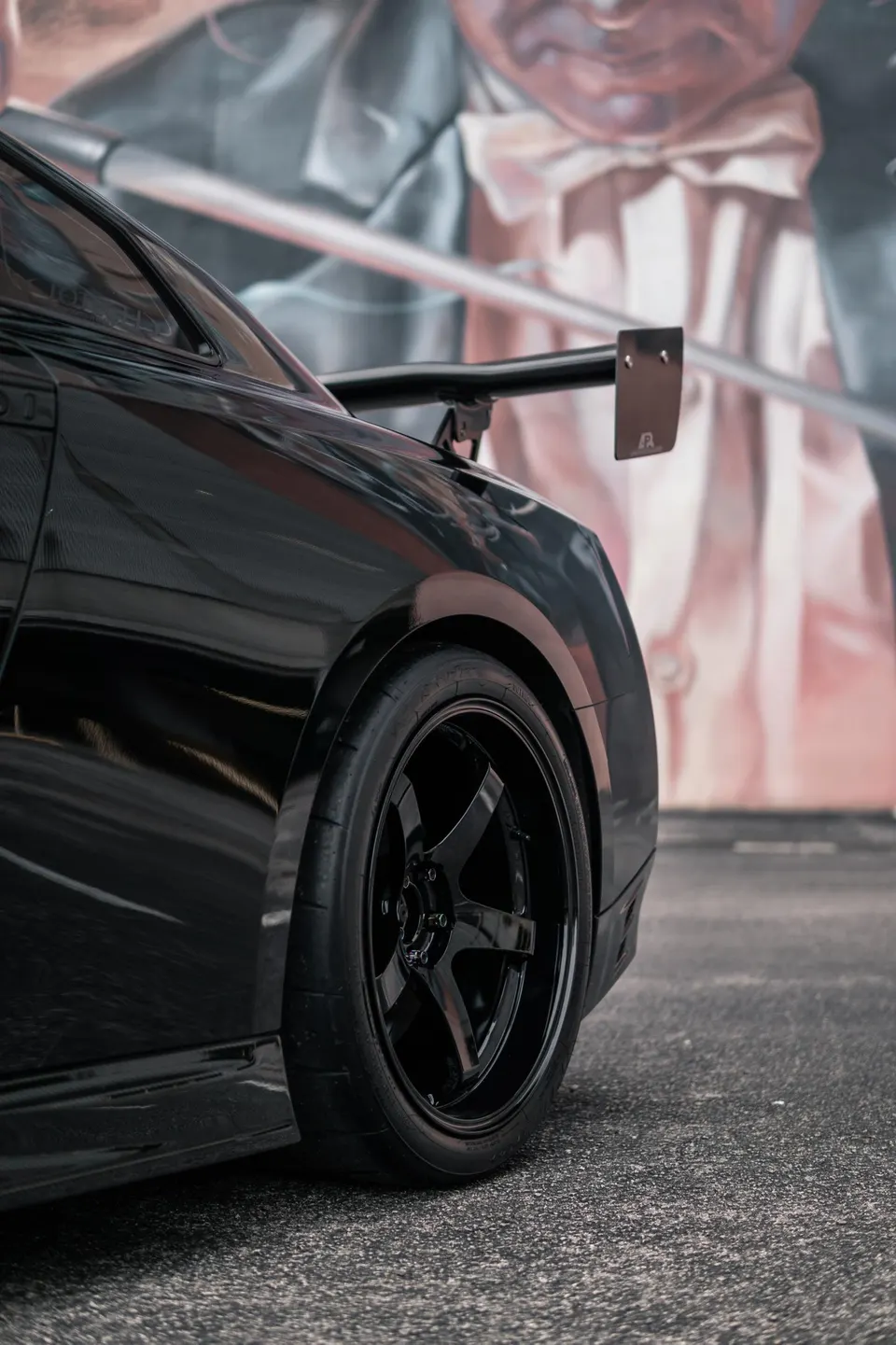 Black sports car with aftermarket wheels, performance tires, and a large rear wing parked in front of a colorful mural.