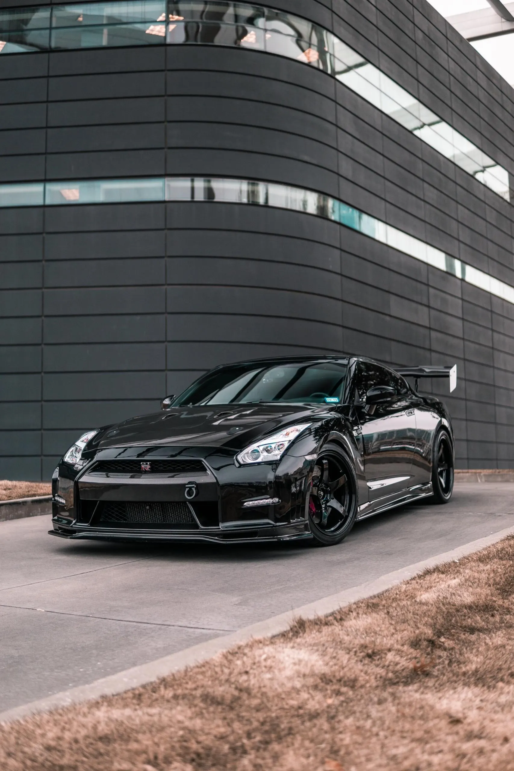 A black Nissan GT-R with aftermarket wheels and a large rear wing parked in front of a modern dark building.
