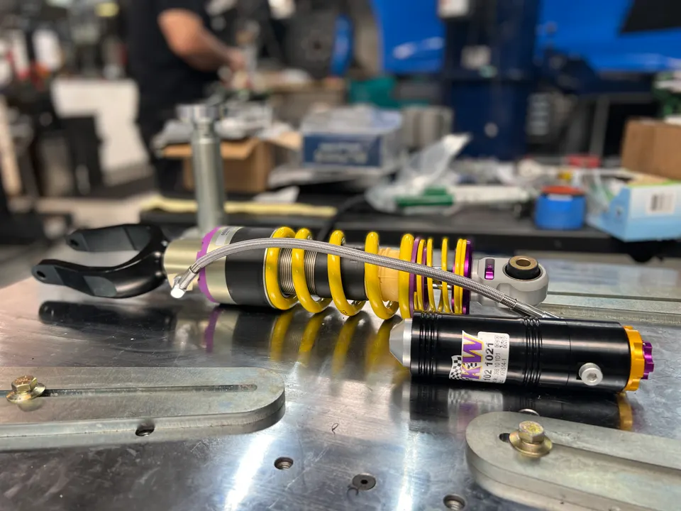 A KW coilover suspension system with an external reservoir on a metal workbench in a performance tuning shop.