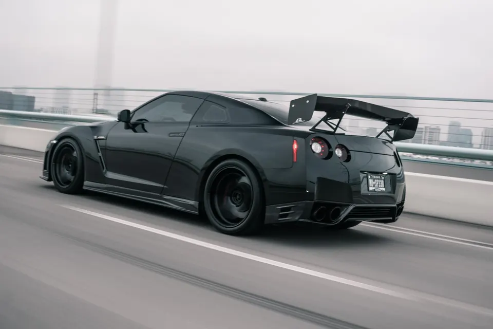 A black Nissan GT-R with an aftermarket rear wing and diffuser drives on a bridge.