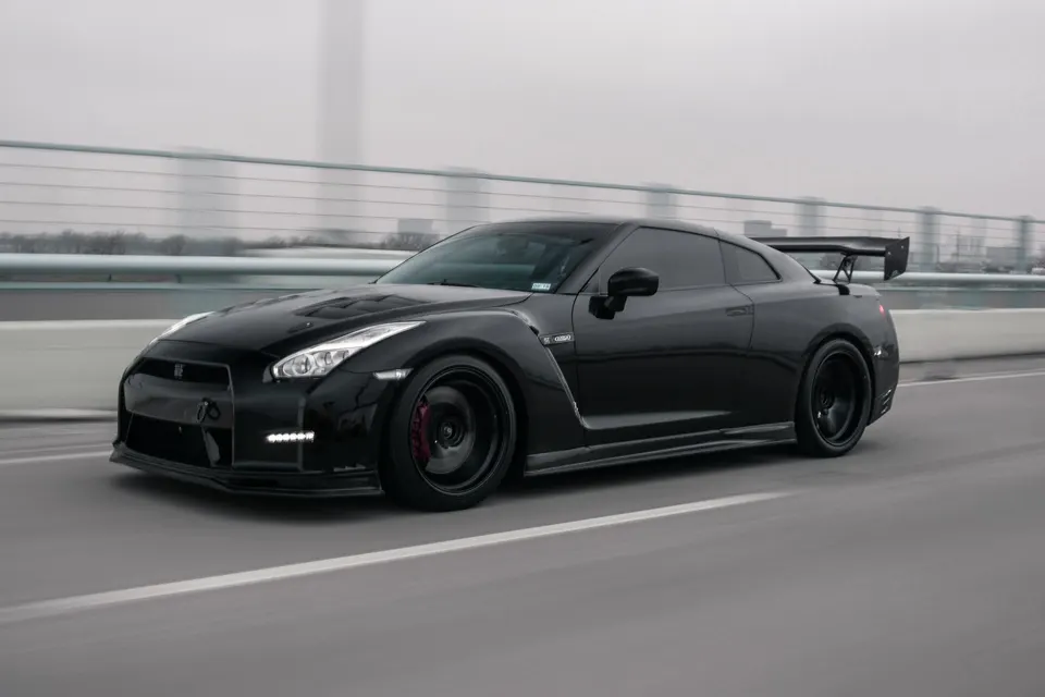 Black Nissan GT-R with aftermarket wheels, front splitter, and large rear wing driving on a bridge.