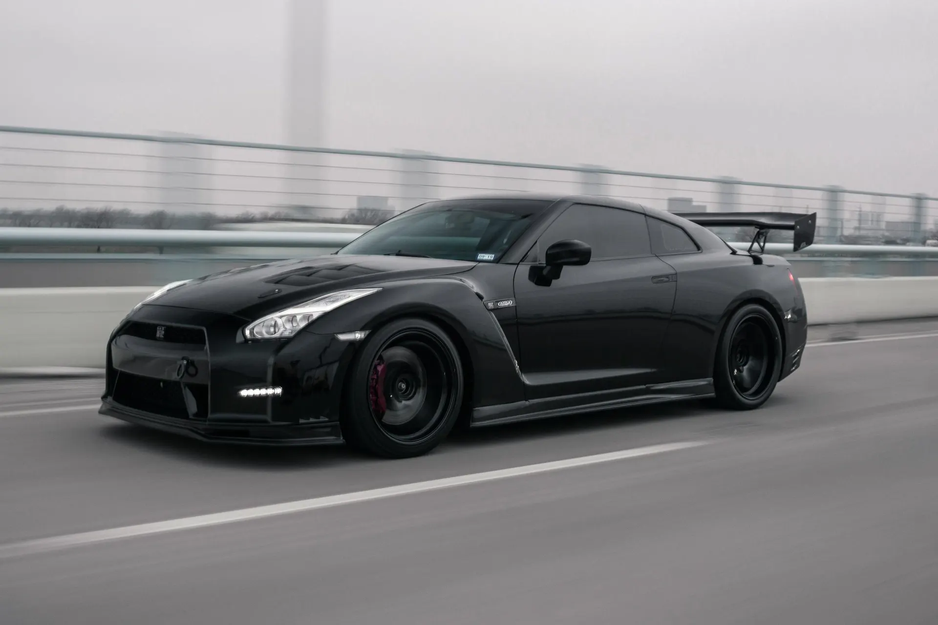 Black Nissan GT-R with aftermarket wheels, front splitter, and large rear wing driving on a bridge.