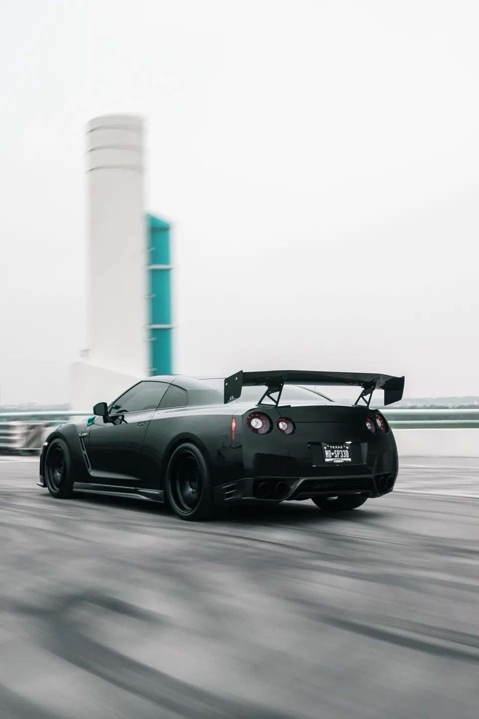 Black Nissan GT-R with a widebody kit and large aftermarket wing in a dynamic roller shot on a bridge.