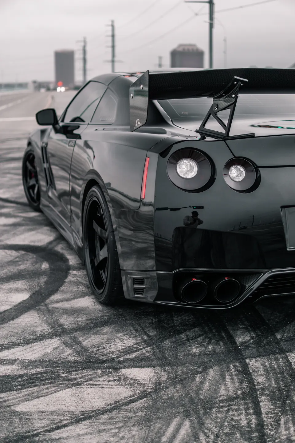 A black Nissan GT-R with a large carbon fiber wing and aftermarket exhaust, parked on a street with tire marks.