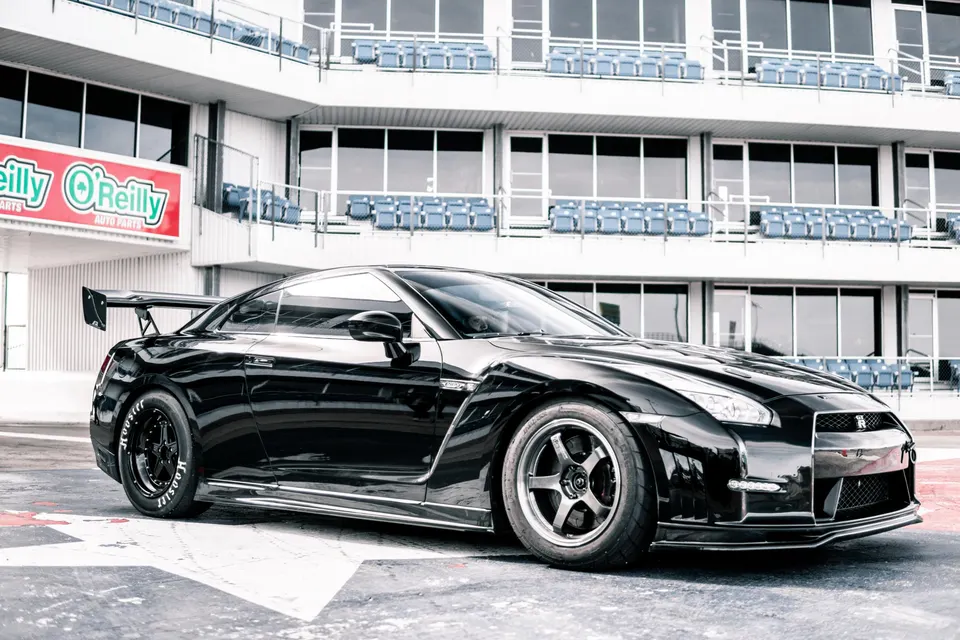 Black Nissan GT-R with aftermarket wheels and a large rear wing parked trackside in front of grandstands.
