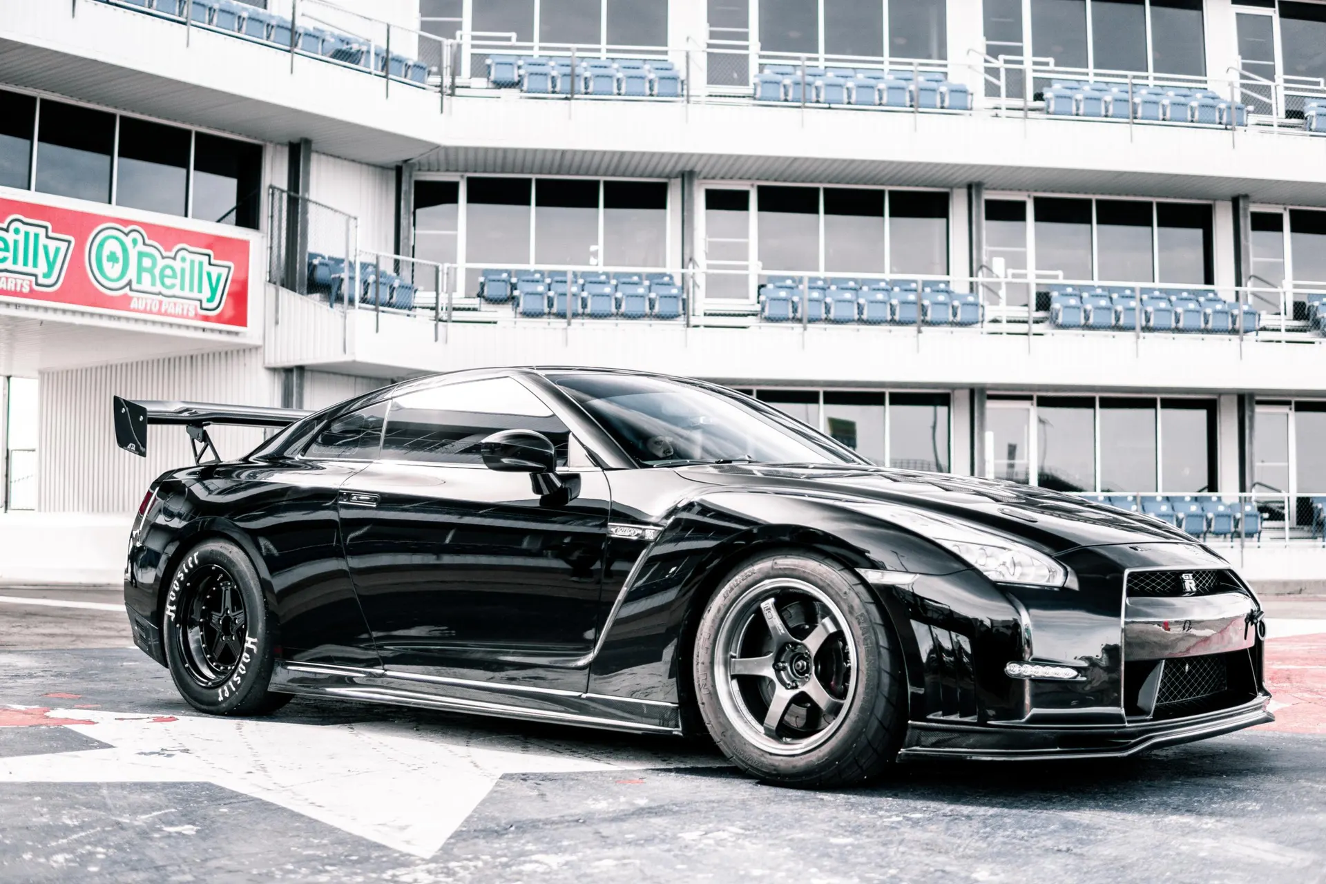 Black Nissan GT-R with aftermarket wheels and a large rear wing parked trackside in front of grandstands.