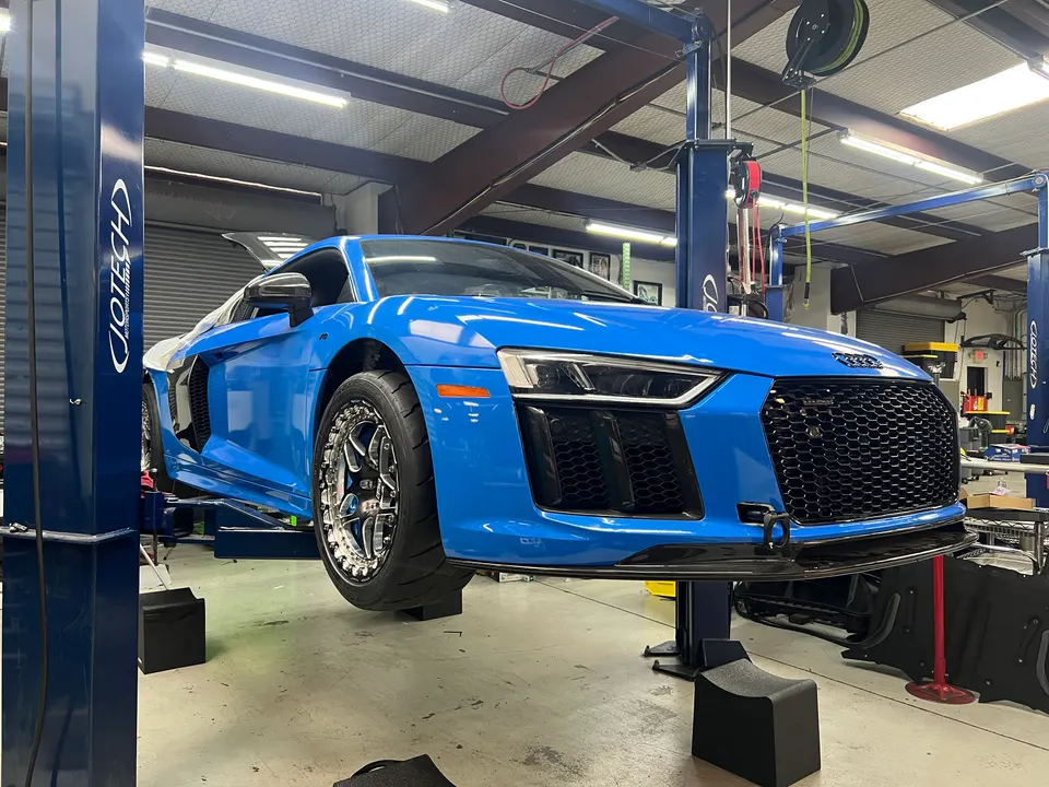 A blue Audi R8 supercar on a lift in a performance tuning shop, showcasing aftermarket wheels and front fascia.