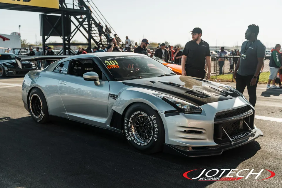 A silver Nissan GT-R with a carbon fiber hood and Hoosier drag tires at a drag strip, with spectators in the background.