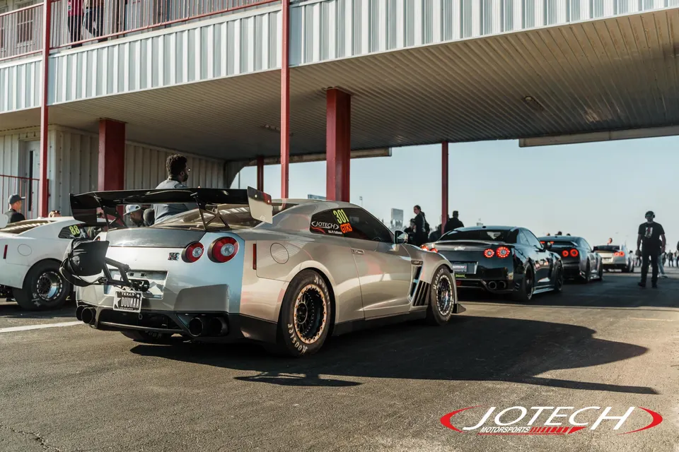 A silver Nissan GT-R with a large rear wing and drag wheels in the staging lanes at a drag strip, with other cars behind it.