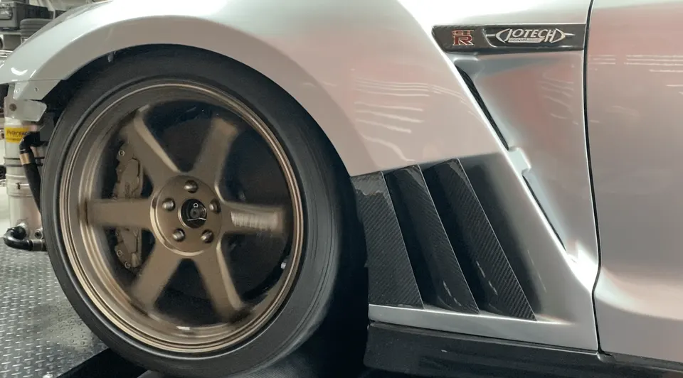 A silver Nissan GT-R on a dyno, showing the spinning bronze wheel, brake caliper, and carbon fiber fender vent.