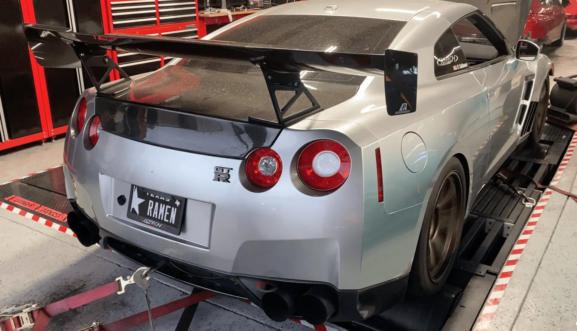A silver Nissan GT-R with a large carbon fiber wing strapped to a dyno at Jotech Motorsports, ready for a power run.