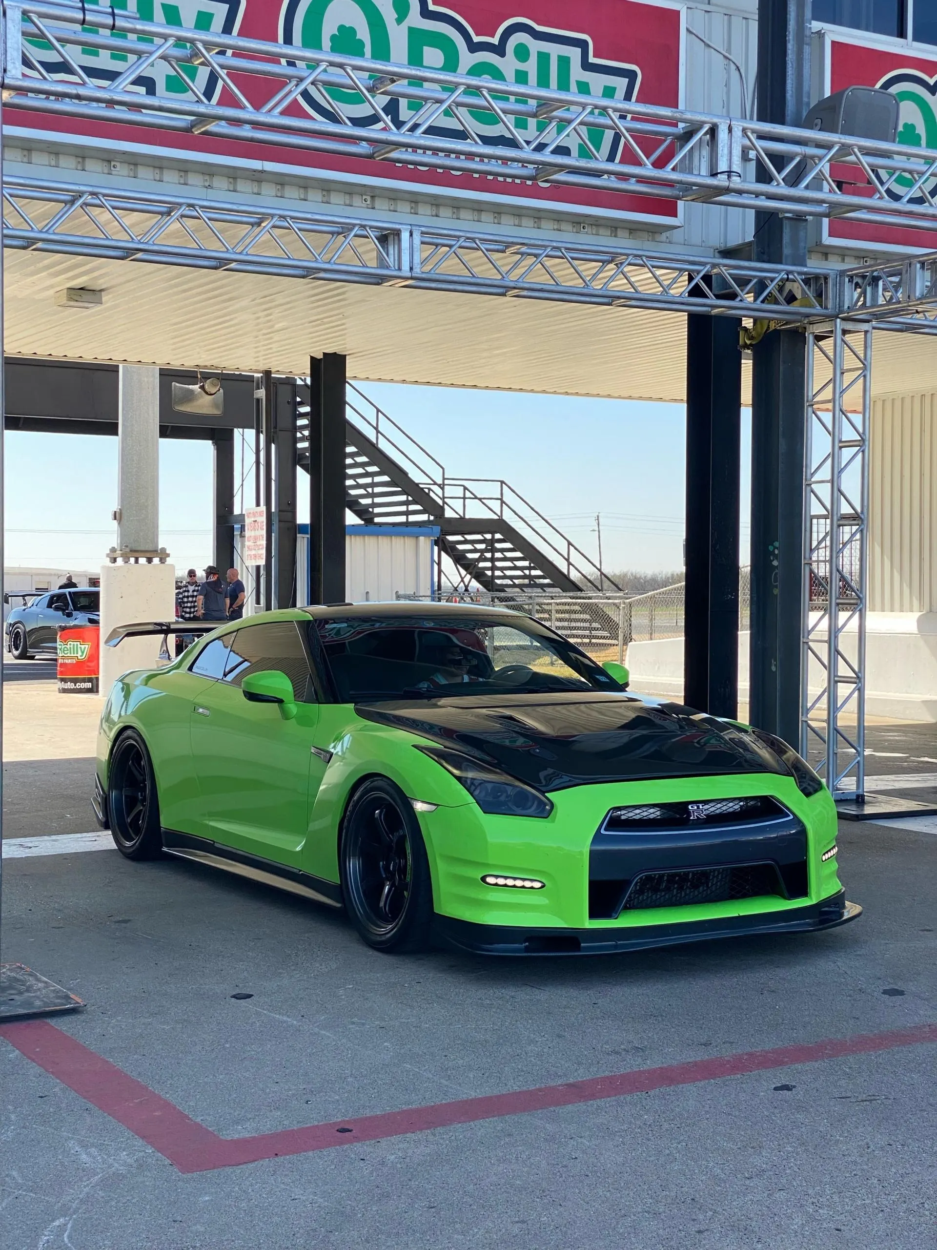 Bright green Nissan GT-R with a black carbon fiber hood and aftermarket wheels parked at a race track.